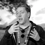 Imagem em preto e branco de Julian LeFay, desenvolvedor de jogos, gesticulando enquanto fala, com cenário natural desfocado ao fundo