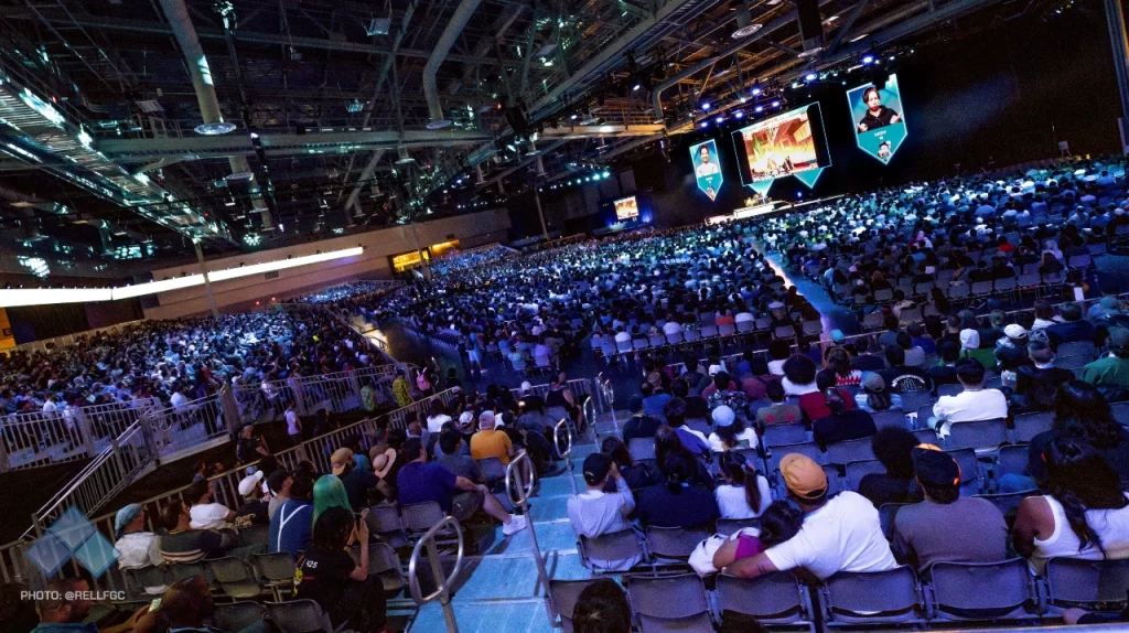 Arena completamente lotada com milhares de espectadores assistindo a uma partida de jogos de luta durante a EVO 2024, em Las Vegas. A cena mostra o palco principal com telões gigantes transmitindo a competição ao vivo.