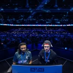 Dois jogadores profissionais de jogos de luta competem em um palco iluminado em azul, diante de uma multidão de espectadores durante a EVO 2024 em Las Vegas. Ambos estão focados nas telas com headsets e crachás de identificação.