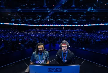 Dois jogadores profissionais de jogos de luta competem em um palco iluminado em azul, diante de uma multidão de espectadores durante a EVO 2024 em Las Vegas. Ambos estão focados nas telas com headsets e crachás de identificação.