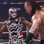 Dois lutadores se encaram no ringue durante uma luta em WWE 2K25, com o logo de WrestleMania e o público ao fundo. Um deles veste uma máscara com pintura de caveira, enquanto o outro exibe tatuagens detalhadas nas costas.