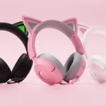 Três headsets gamer com orelhas de gato alinhados sobre fundo rosa: um preto com detalhes verdes, um rosa com cinza e um branco com detalhes rosa.