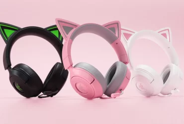 Três headsets gamer com orelhas de gato alinhados sobre fundo rosa: um preto com detalhes verdes, um rosa com cinza e um branco com detalhes rosa.