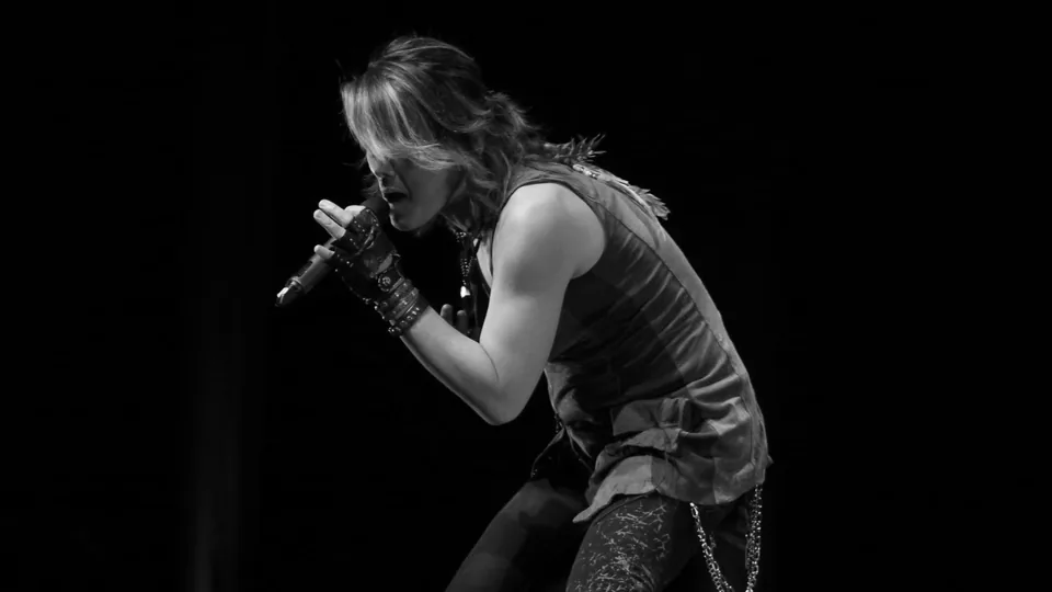 Cantor japonês se apresenta no palco segurando um microfone com as duas mãos, em foto em preto e branco.