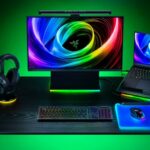 Setup gamer da Razer com teclado mecânico iluminado, mouse sobre mousepad RGB, headset em suporte iluminado, monitor e notebook exibindo papel de parede com cores vibrantes.
