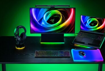 Setup gamer da Razer com teclado mecânico iluminado, mouse sobre mousepad RGB, headset em suporte iluminado, monitor e notebook exibindo papel de parede com cores vibrantes.