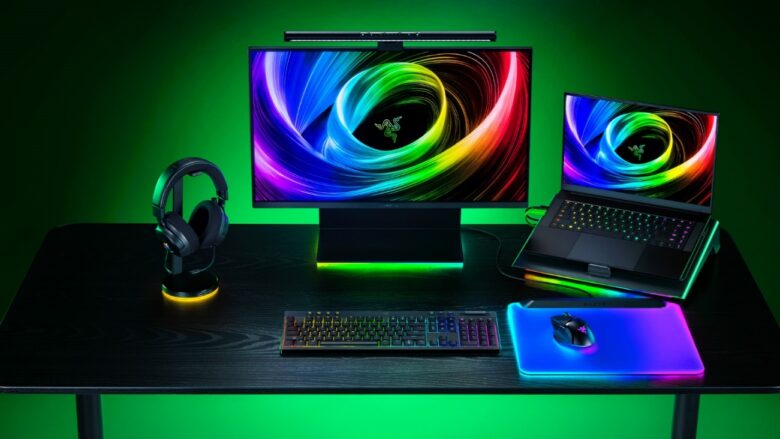 Setup gamer da Razer com teclado mecânico iluminado, mouse sobre mousepad RGB, headset em suporte iluminado, monitor e notebook exibindo papel de parede com cores vibrantes.