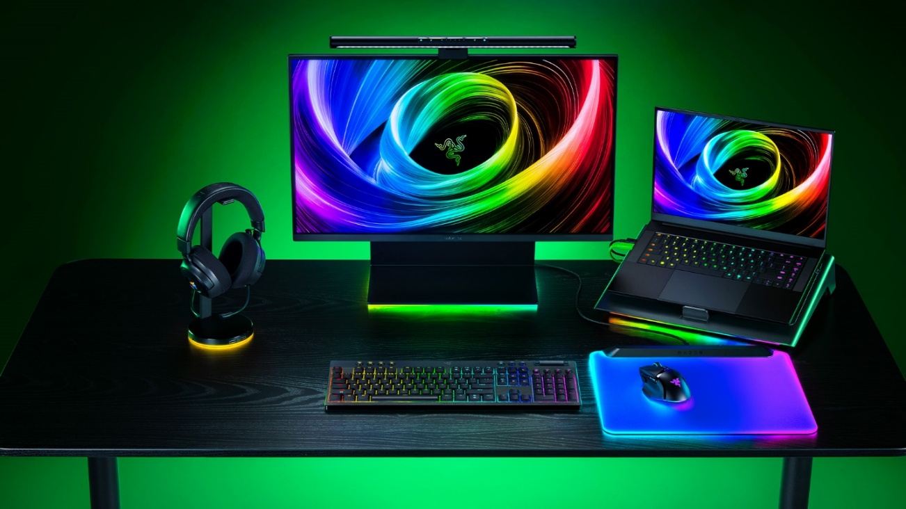Setup gamer da Razer com teclado mecânico iluminado, mouse sobre mousepad RGB, headset em suporte iluminado, monitor e notebook exibindo papel de parede com cores vibrantes.