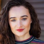 Retrato da atriz Kelley Mack sorrindo levemente, com cabelos castanhos cacheados soltos, batom vermelho e uma blusa listrada colorida. Ela está em ambiente externo com fundo desfocado.