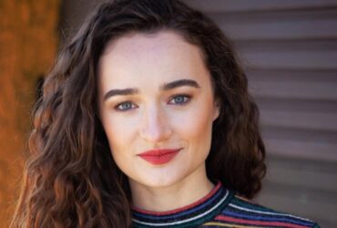 Retrato da atriz Kelley Mack sorrindo levemente, com cabelos castanhos cacheados soltos, batom vermelho e uma blusa listrada colorida. Ela está em ambiente externo com fundo desfocado.