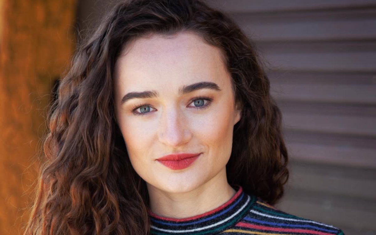 Retrato da atriz Kelley Mack sorrindo levemente, com cabelos castanhos cacheados soltos, batom vermelho e uma blusa listrada colorida. Ela está em ambiente externo com fundo desfocado.