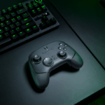 Controle sem fio Razer Wolverine V3 Pro 8K PC sobre uma mesa gamer preta, ao lado de um teclado mecânico iluminado com luz verde e um mouse, em ambiente com iluminação de tom esverdeado.