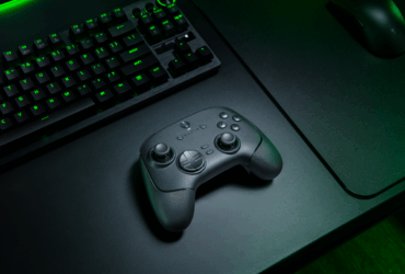 Controle sem fio Razer Wolverine V3 Pro 8K PC sobre uma mesa gamer preta, ao lado de um teclado mecânico iluminado com luz verde e um mouse, em ambiente com iluminação de tom esverdeado.