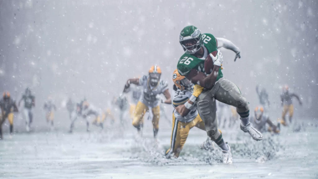 Running back dos Philadelphia Eagles, camisa 26, corre com a bola durante uma forte nevasca enquanto é perseguido e atingido por defensores do Green Bay Packers; jogadores ao fundo aparecem desfocados pelo vento e pela neve.