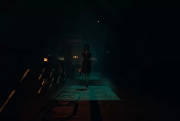 Figura sombria de uma mulher com vestido escuro e cabelo cobrindo parte do rosto caminha em um corredor escuro e empoeirado, segurando um urso de pelúcia em uma das mãos. A cena tem iluminação baixa e atmosfera de terror.