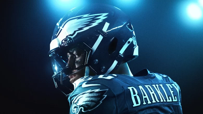 Close-up de um atleta da NFL com uniforme dos Philadelphia Eagles, visto de perfil com capacete preto e logo da águia, pintura no rosto e iluminação azul intensa ao fundo.