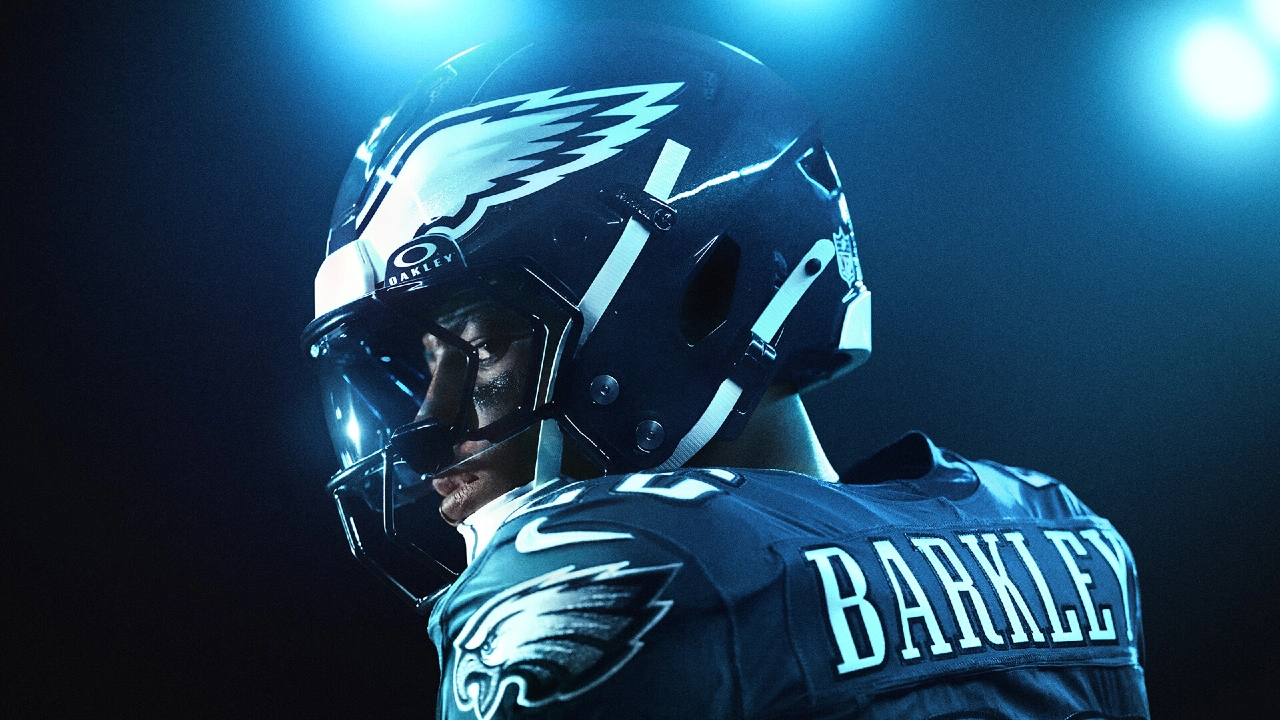 Close-up de um atleta da NFL com uniforme dos Philadelphia Eagles, visto de perfil com capacete preto e logo da águia, pintura no rosto e iluminação azul intensa ao fundo.