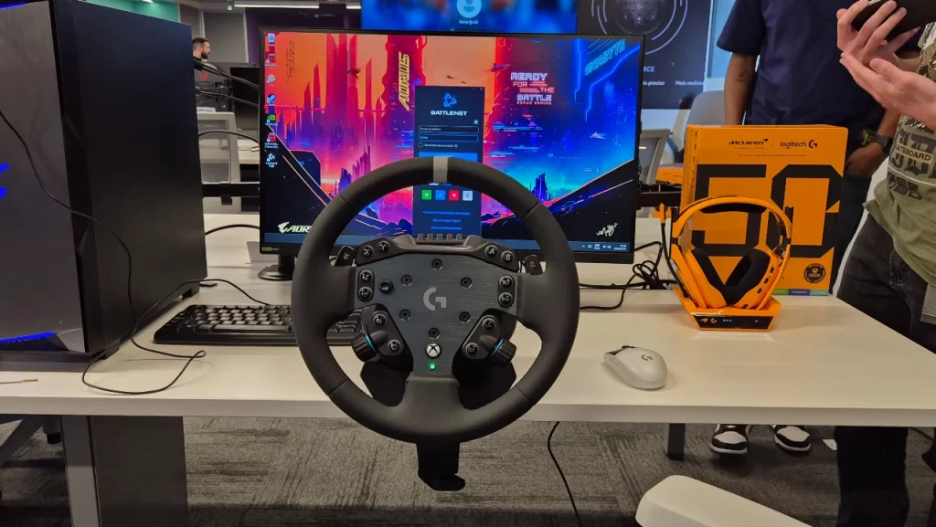Volante Logitech G RS50 conectado a um PC gamer, acompanhado do headset Astro A50 X McLaren em cor laranja, exibido em mesa de demonstração durante evento de lançamento.