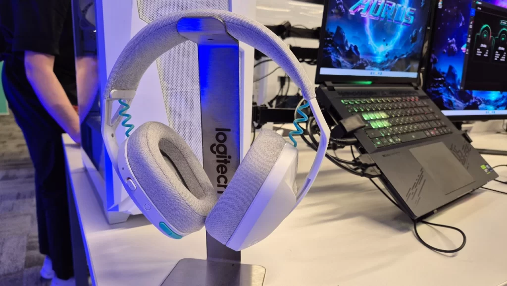 Headset sem fio Logitech G321 LIGHTSPEED na cor branca, exibido em suporte com logo da Logitech, em ambiente de demonstração com PCs e periféricos gamers ao fundo.