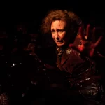 Cena de Invocação do Mal 4: O Último Ritual mostra Lorraine Warren, interpretada por Vera Farmiga, ferida e coberta de sangue, com expressão de medo e a mão estendida em um ambiente escuro e tenso.