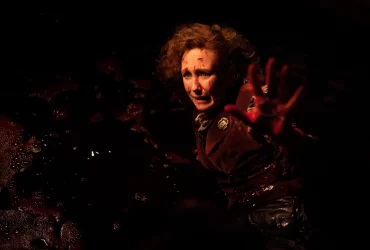 Cena de Invocação do Mal 4: O Último Ritual mostra Lorraine Warren, interpretada por Vera Farmiga, ferida e coberta de sangue, com expressão de medo e a mão estendida em um ambiente escuro e tenso.