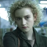 Cena de uma personagem jovem com expressão séria, cabelos loiros cacheados e curtos, em ambiente interno iluminado.