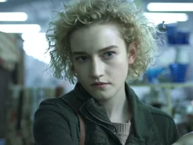 Cena de uma personagem jovem com expressão séria, cabelos loiros cacheados e curtos, em ambiente interno iluminado.