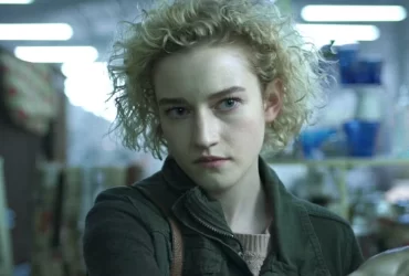 Cena de uma personagem jovem com expressão séria, cabelos loiros cacheados e curtos, em ambiente interno iluminado.