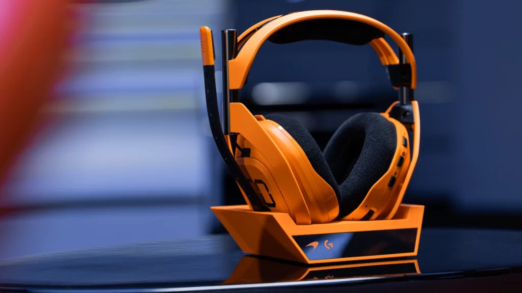 Headset sem fio Astro A50 X McLaren Racing Edition em laranja Papaya, posicionado sobre sua base de carregamento, com fundo desfocado em tom azul.