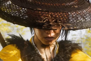 Personagem samurai do jogo Ghost of Yotei para PlayStation 5 em destaque, com chapéu tradicional e cenário de batalha ao fundo.