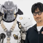 Hideo Kojima posando ao lado de uma estátua futurista em armadura branca com detalhes dourados e capacete transparente.