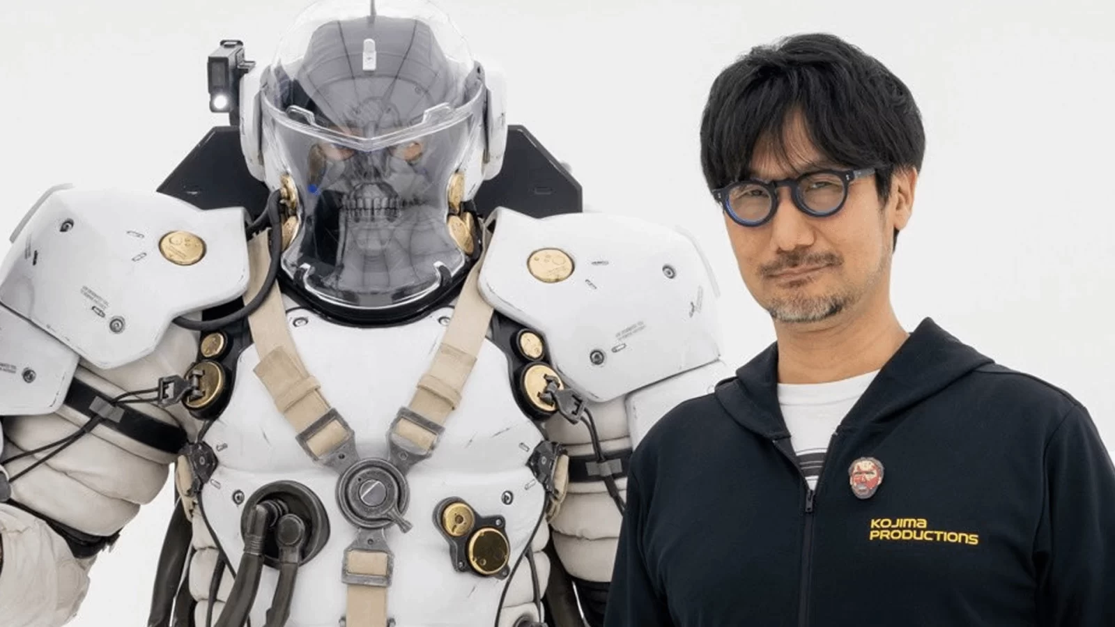 Hideo Kojima posando ao lado de uma estátua futurista em armadura branca com detalhes dourados e capacete transparente.