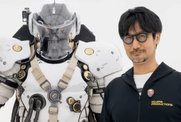 Hideo Kojima posando ao lado de uma estátua futurista em armadura branca com detalhes dourados e capacete transparente.