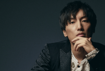 Retrato profissional em estúdio de Hiroyuki Sawano, compositor japonês de trilhas sonoras de anime. Homem asiático de cabelos pretos médios, vestindo blazer escuro sobre camisa clara, em pose pensativa com mão no queixo, contra fundo escuro com iluminação dramática lateral.