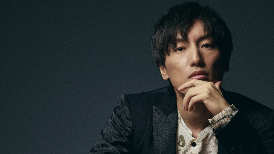 Retrato profissional em estúdio de Hiroyuki Sawano, compositor japonês de trilhas sonoras de anime. Homem asiático de cabelos pretos médios, vestindo blazer escuro sobre camisa clara, em pose pensativa com mão no queixo, contra fundo escuro com iluminação dramática lateral.