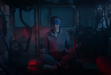 Personagem de Stranger Things 5 senta no chão com os olhos vendados em um ambiente escuro tomado por raízes e luzes vermelhas do Mundo Invertido.