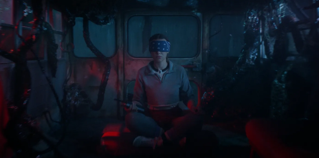 Personagem de Stranger Things 5 senta no chão com os olhos vendados em um ambiente escuro tomado por raízes e luzes vermelhas do Mundo Invertido.