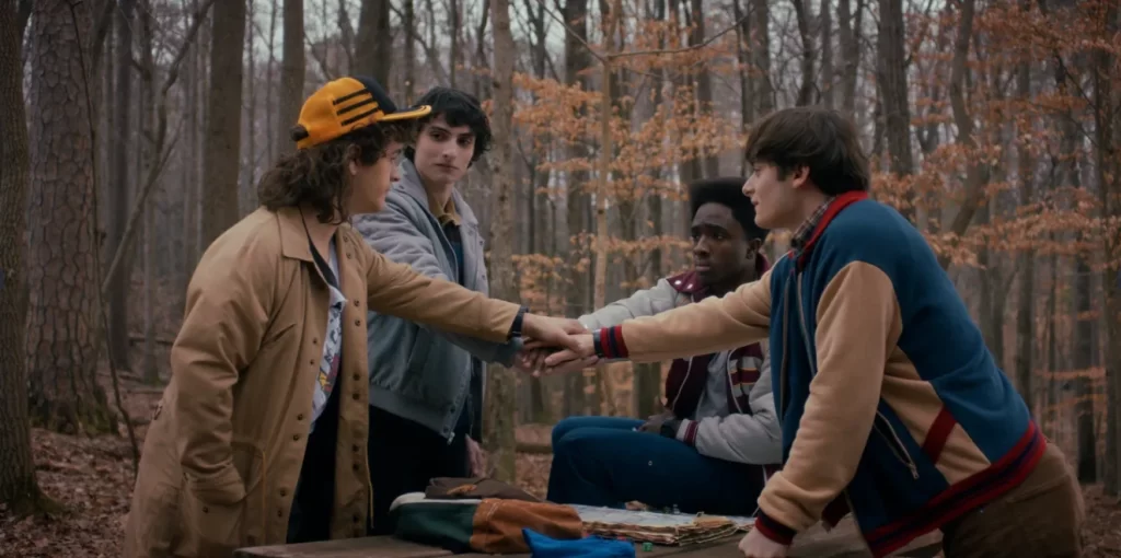 Quatro amigos de Stranger Things estendem as mãos uns aos outros em um gesto de união no meio de uma floresta durante o outono.