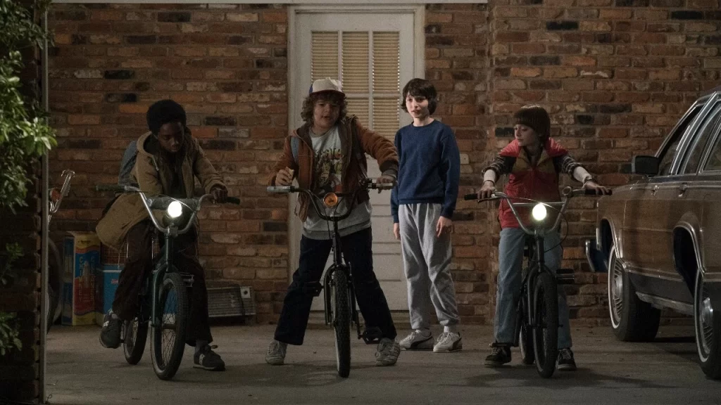 Quatro garotos com bicicletas parados em uma garagem à noite, personagens da 1ª temporada de Stranger Things, em Hawkins.