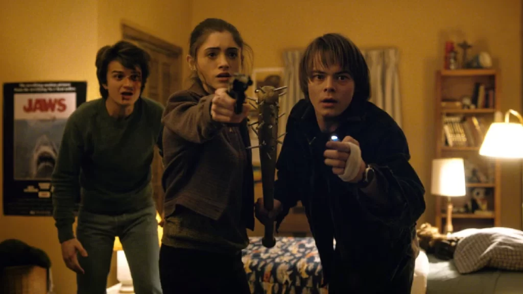 Três jovens em um quarto apontam armas improvisadas e uma lanterna, cena tensa da 1ª temporada de Stranger Things.