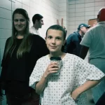 Pessoa caracterizada com roupa hospitalar segura um copo em bastidores da 1ª temporada de Stranger Things, com equipe ao fundo.