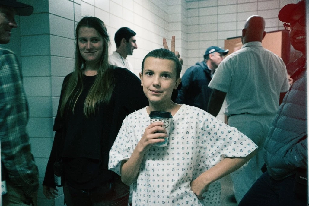 Pessoa caracterizada com roupa hospitalar segura um copo em bastidores da 1ª temporada de Stranger Things, com equipe ao fundo.
