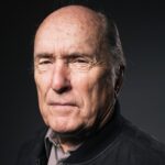 Retrato de Robert Duvall com expressão séria, usando jaqueta escura e camisa, contra fundo neutro escuro.