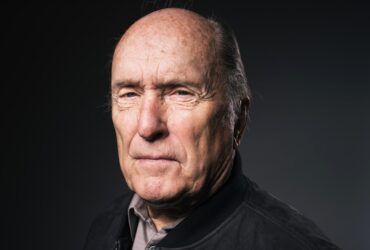 Retrato de Robert Duvall com expressão séria, usando jaqueta escura e camisa, contra fundo neutro escuro.