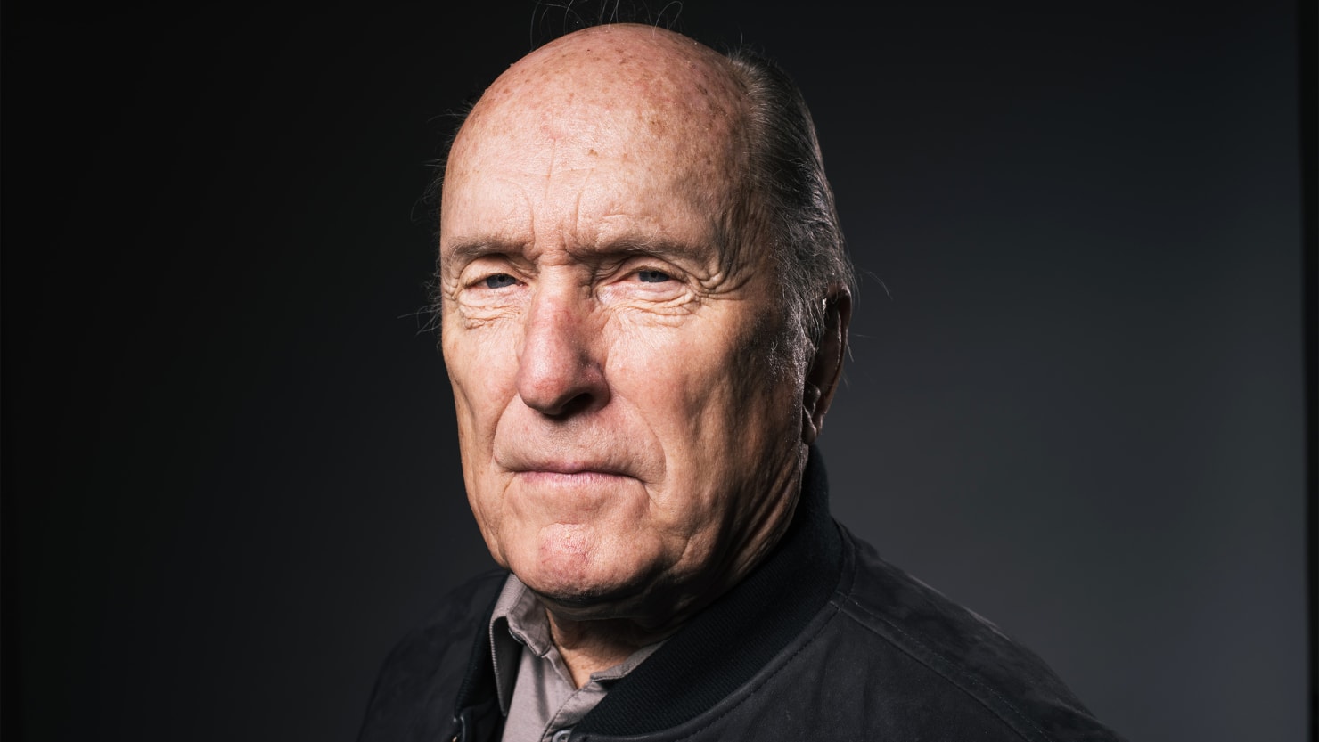 Retrato de Robert Duvall com expressão séria, usando jaqueta escura e camisa, contra fundo neutro escuro.