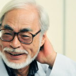 Retrato de Hayao Miyazaki sorrindo, usando óculos, com cabelo branco e barba, em fundo claro.