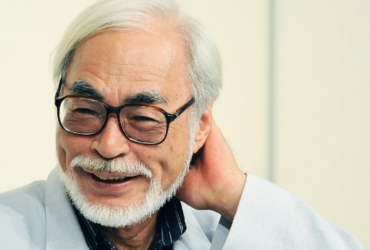 Retrato de Hayao Miyazaki sorrindo, usando óculos, com cabelo branco e barba, em fundo claro.