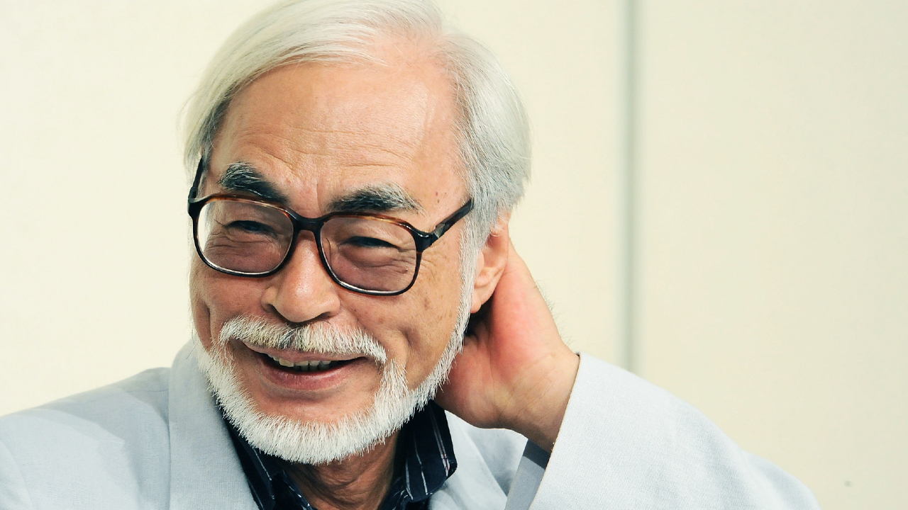 Retrato de Hayao Miyazaki sorrindo, usando óculos, com cabelo branco e barba, em fundo claro.