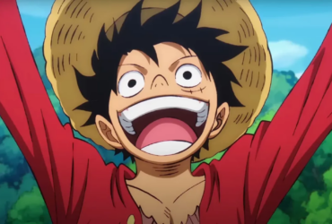 Monkey D. Luffy, personagem de One Piece, aparece sorrindo com a boca aberta e os braços erguidos, usando chapéu de palha e camisa vermelha, com céu azul e vegetação ao fundo.
