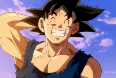 Goku, personagem de Dragon Ball, aparece sorrindo com os olhos fechados e os dentes à mostra, com a mão atrás da cabeça, usando roupa rasgada, com céu azul e nuvens ao fundo.